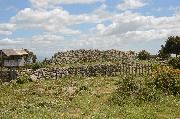 Die zum Plateau-Inneren orientierte, leicht eingewölbte Seite des Nuraghen in geschlossener Mauerung aus besonders großen Basaltblöcken hinter der Mauer um das Gelände. Davor vorne links die Infotafel zum Nuraghen und seinem Dorf.